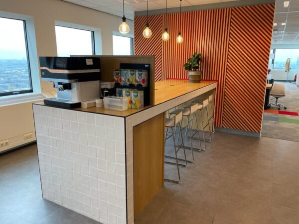 Moderne koffiehoek met een lange houten tafel, barkrukken en een koffieautomaat op het Johanna Westerdijkplein.